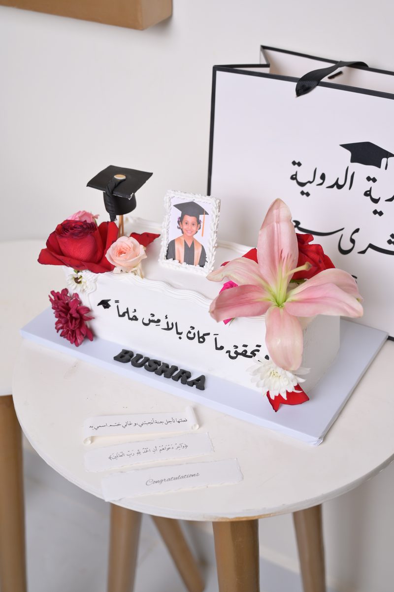 كيكة تخرج بيضاء عليها صورة الخريجة وقبعة تخرج وورود وزنبق، ومكتوب عليها: تحقق ما كان بالأمس حلماً