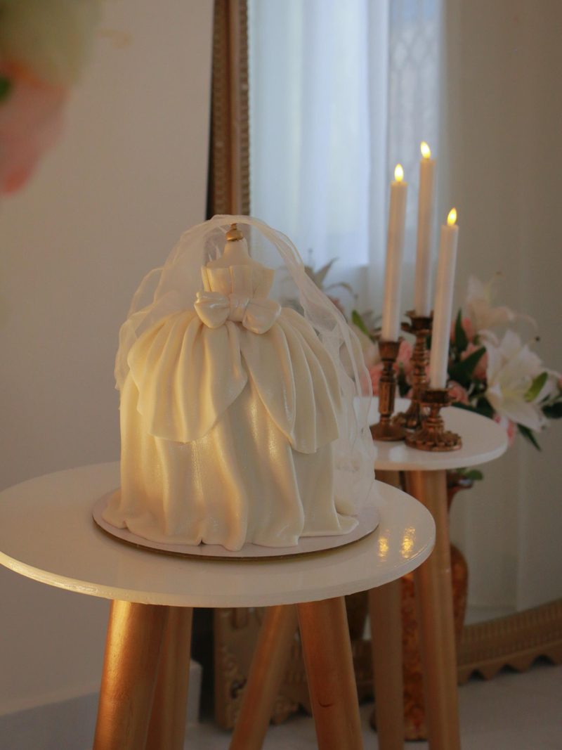 كيكة على شكل فستان عروس أبيض بفيونكة وطرحة شفافة، وبجانبها شموع